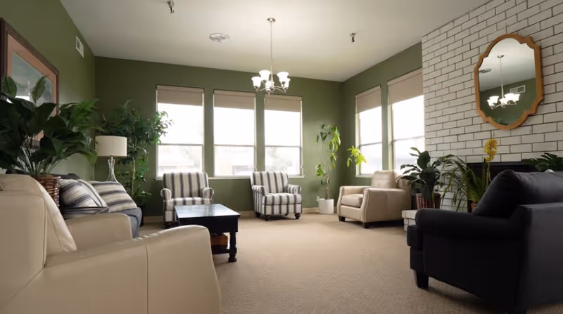 A bright and spacious living room with green walls and beige carpet. The room features multiple armchairs including striped and solid-colored ones, a dark wooden coffee table, several potted plants, a white brick fireplace with a wooden-framed mirror above it, and three large windows with blinds letting in natural light.