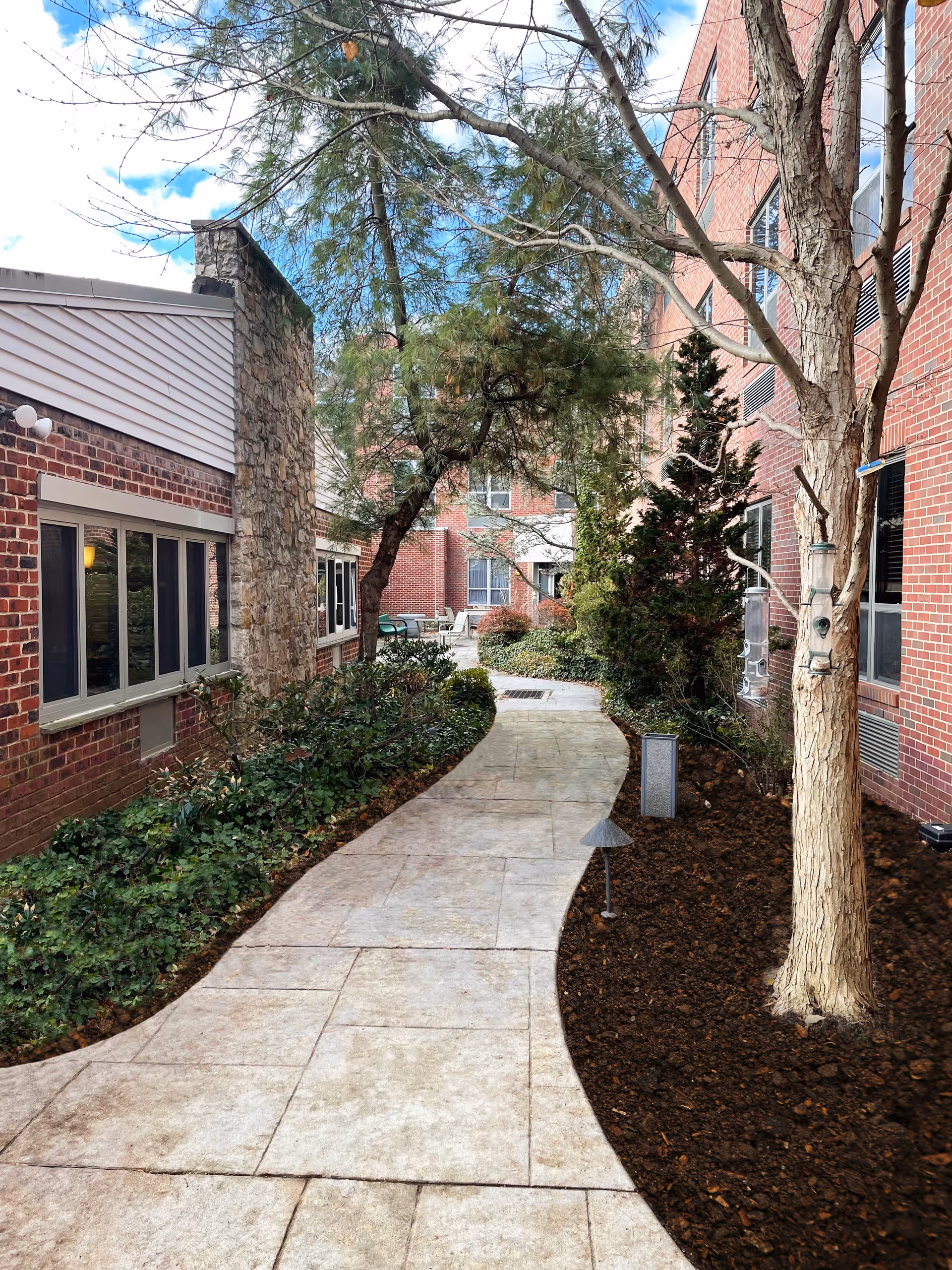 Curved paved walkway between two brick buildings with windows, surrounded by landscaped greenery including bushes, trees, and mulch beds. Outdoor seating is visible in the distance under a partly cloudy sky.