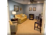 A small living room with beige carpet, light blue walls, and a ceiling fan. The room contains a yellow sofa with decorative pillows, a patterned armchair, a wooden side table with a lamp, a small desk with a chair, and two additional wooden chairs. Three framed pictures hang on the wall above the desk.