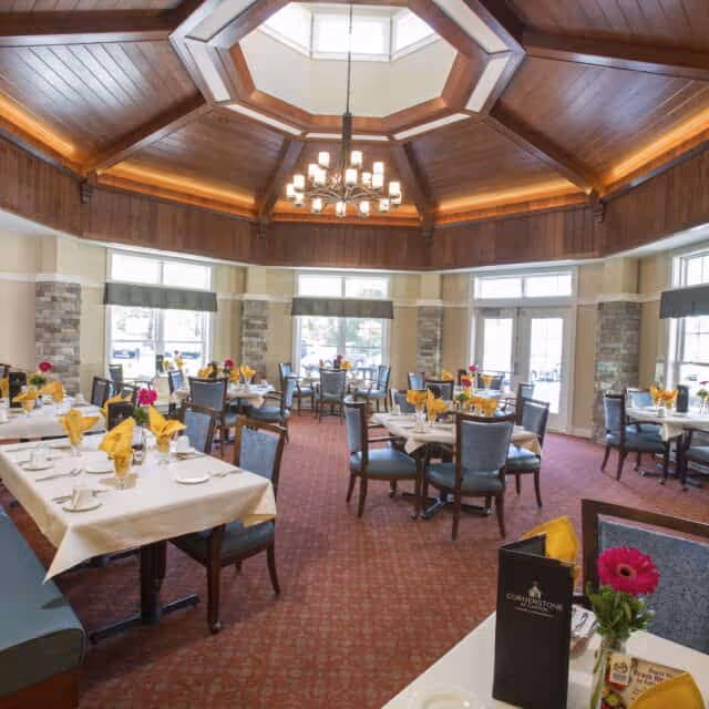 A spacious dining room with multiple tables set with white tablecloths, yellow folded napkins, and glassware. The room features large windows allowing natural light, stone pillars, a red carpet, and a wooden octagonal ceiling with a chandelier hanging in the center.