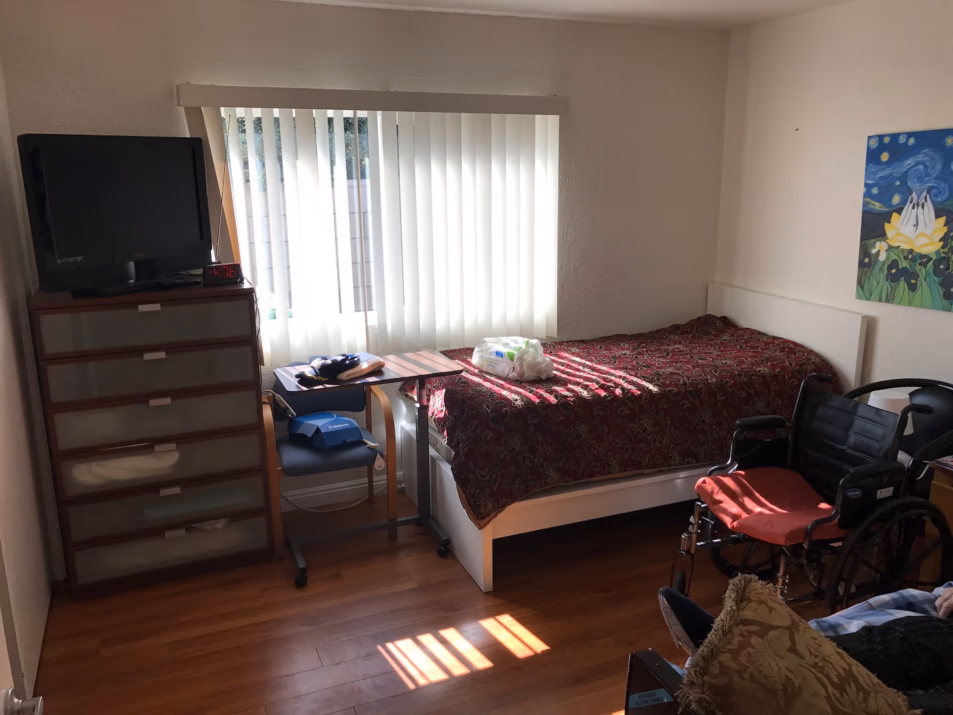 A small bedroom with a single bed covered with a red patterned blanket, a wheelchair with a red cushion, a wooden dresser with a TV on top, a chair with a small table attached, and a window with vertical blinds letting in sunlight. There is a colorful painting on the wall and wooden flooring.