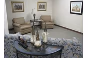 A small sitting area with two beige armchairs and a glass-top side table between them. Behind the armchairs is a plain white wall with two framed pictures. In the foreground, there is a patterned sofa with a decorative console table behind it, holding three ornamental vases.