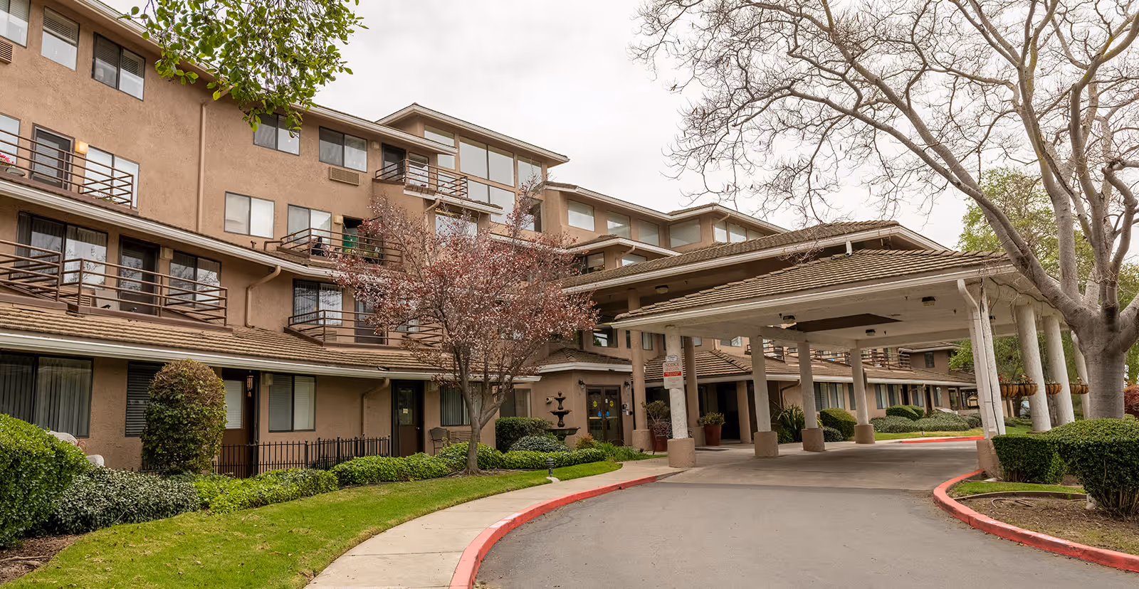 Exterior view of a multi-story retirement community building with balconies, a covered entrance driveway, landscaped greenery, and trees with some leaves missing.
