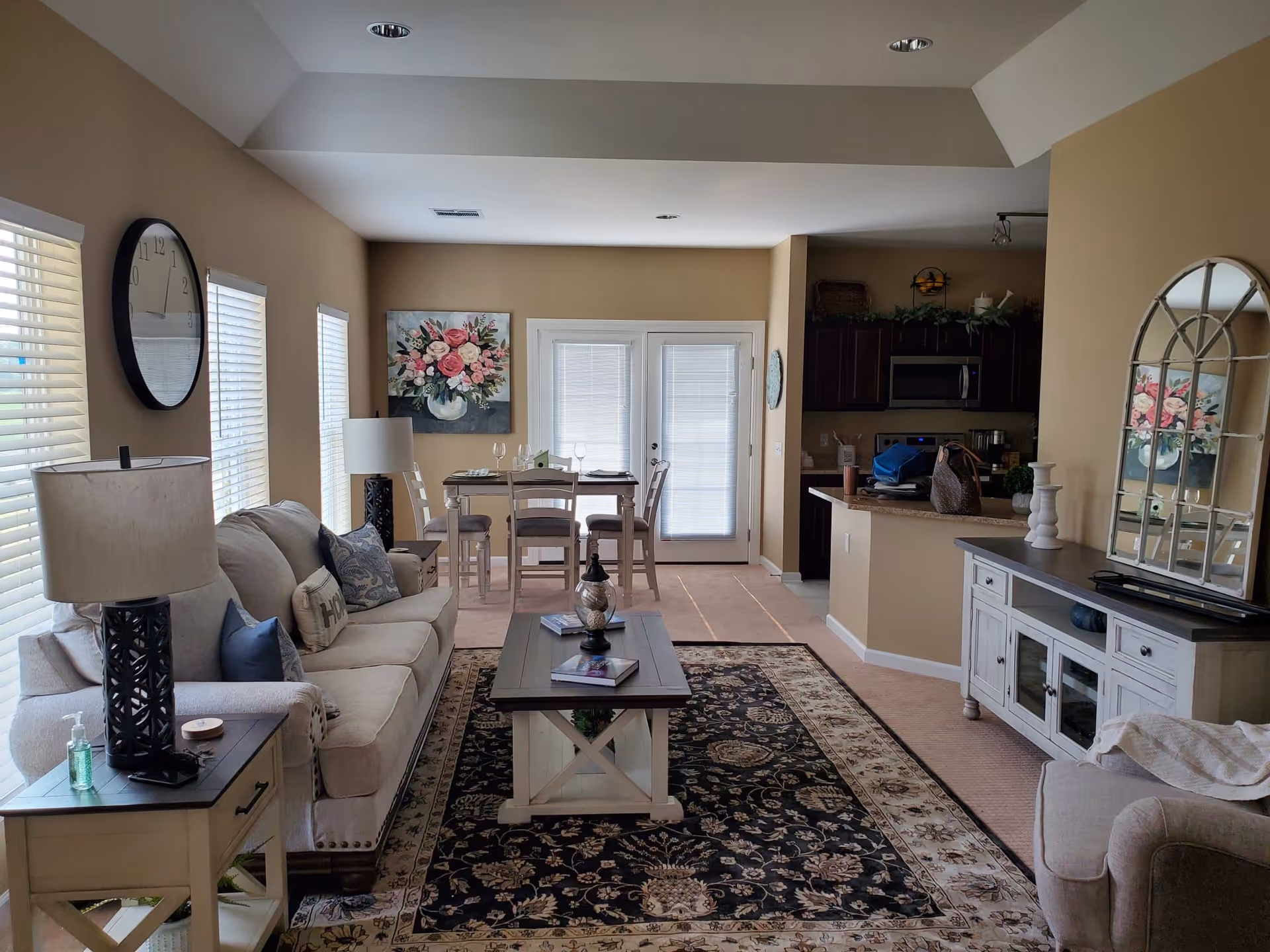 A cozy living room and dining area with beige walls and carpet. The living room features a beige sofa with decorative pillows, a wooden coffee table on a patterned rug, a side table with a lamp, and a large wall clock. The dining area has a wooden table with four chairs, set near double glass doors with blinds. A floral painting hangs on the wall above the dining table. To the right, there is a kitchen area with dark wooden cabinets, a microwave, and a countertop with various items. A large decorative mirror and a white cabinet are also visible.