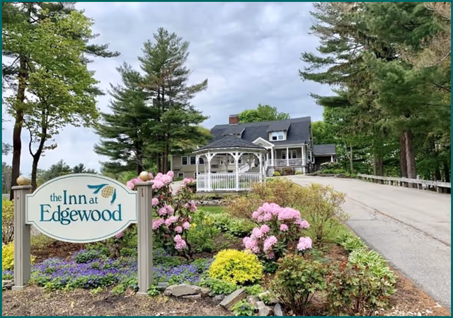 View of The Inn at Edgewood facility showing a large house with a dark roof, surrounded by tall trees and landscaped garden with blooming pink and purple flowers. A white gazebo is visible in front of the house, and a paved driveway leads up to the building. A sign with the facility name is prominently displayed in the foreground.