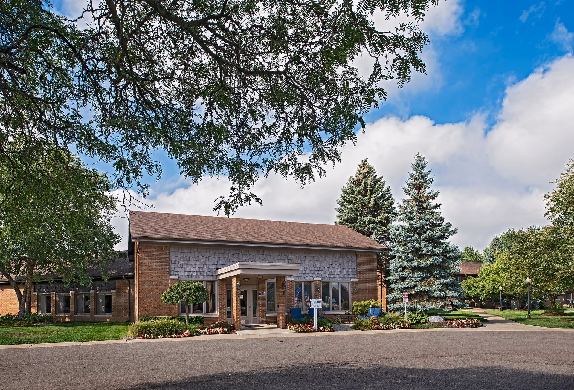 Exterior view of a single-story brick building with a brown roof, surrounded by green trees and landscaped flower beds under a partly cloudy blue sky.