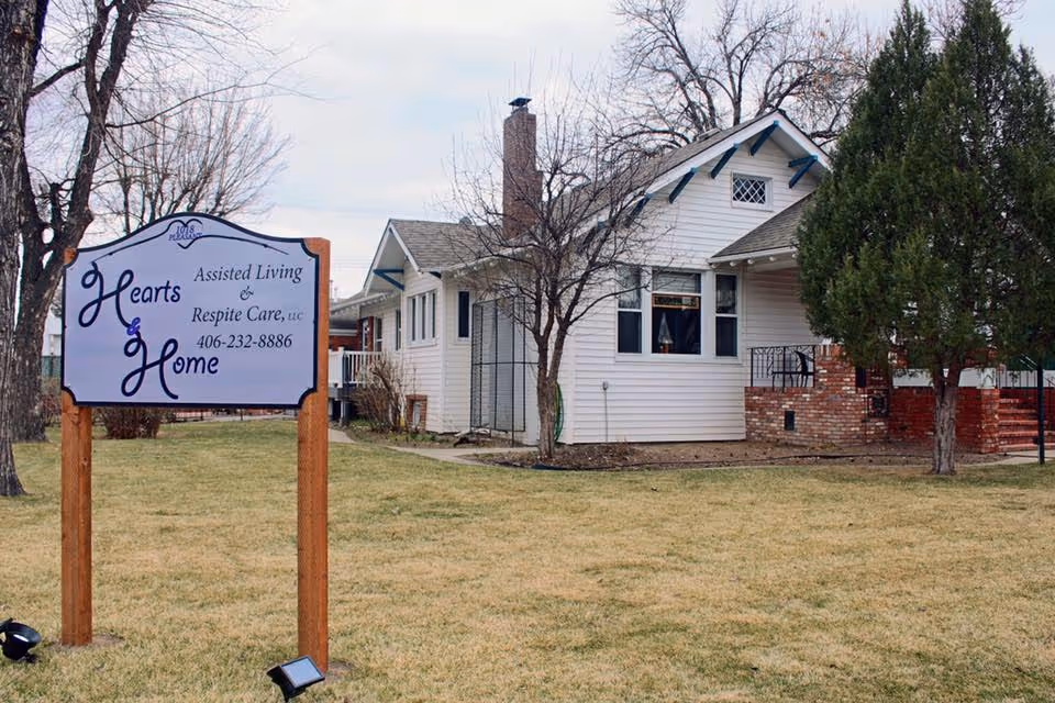 Exterior view of a single-story assisted living facility named Hearts & Home Assisted Living & Respite Care. The building is white with a brick porch and surrounded by a lawn with some trees. A sign with the facility's name and phone number is prominently displayed on the grass near the sidewalk.