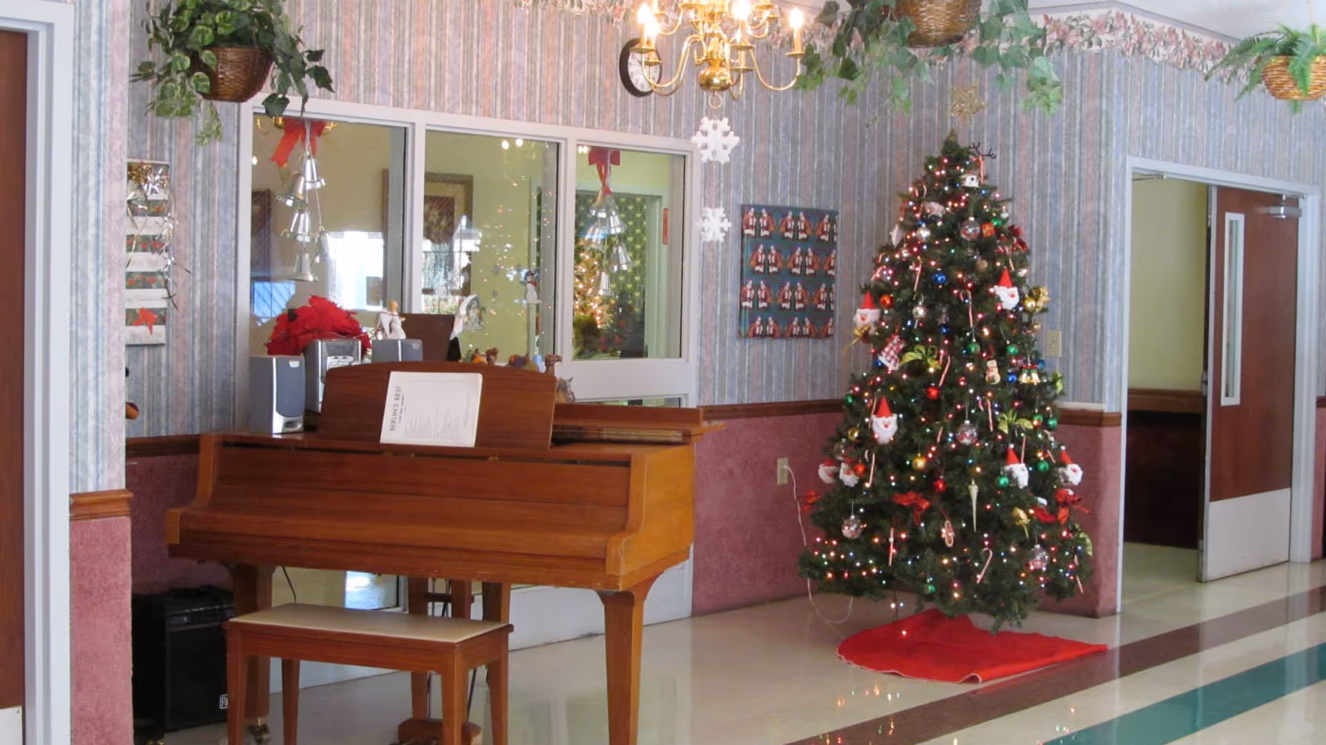 Interior of a senior living facility decorated for Christmas with a decorated Christmas tree, a wooden piano with a bench, hanging plants, and a chandelier. The walls have striped wallpaper with a floral border near the ceiling, and there are windows and doors in the background.
