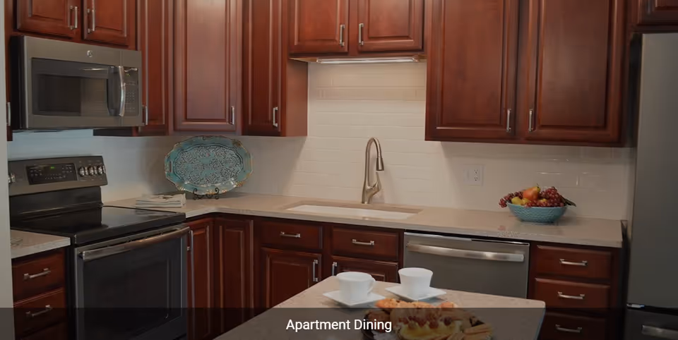 Modern kitchen with dark wooden cabinets, stainless steel appliances including a microwave, stove, dishwasher, and refrigerator. The countertop has a sink with a sleek faucet, a decorative plate, a bowl of fruit, and a small table set with two white cups and plates with pastries.