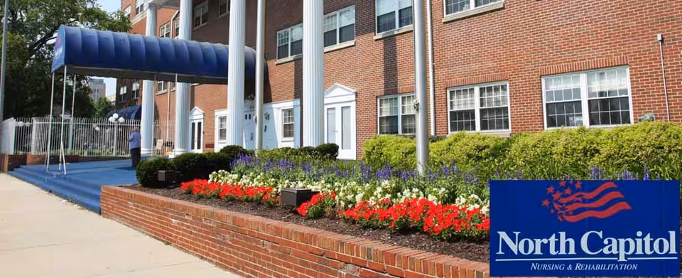 Exterior view of North Capitol Nursing and Rehabilitation building with a blue awning over the entrance, a brick facade, and a landscaped flower bed with red, white, and purple flowers in front.