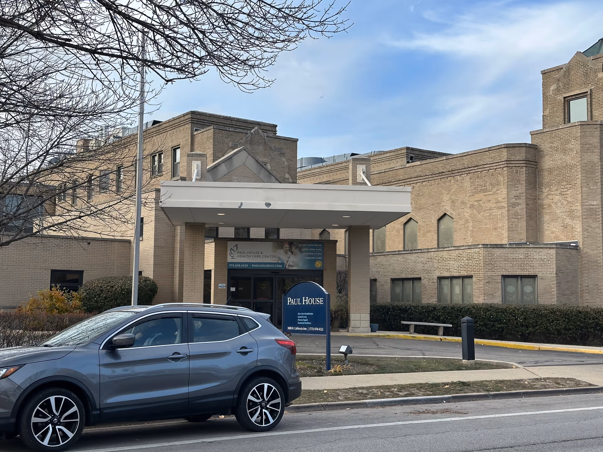 Exterior view of Paul House and Healthcare Center building with a gray SUV parked on the street in front. The building is made of light brown bricks and has a covered entrance with a sign displaying the facility's name and contact information. Leafless tree branches are visible in the foreground under a partly cloudy sky.