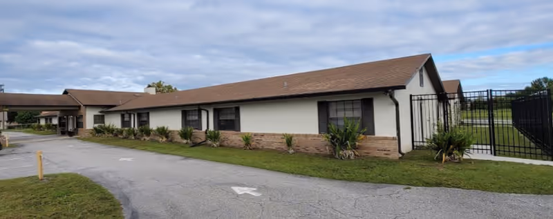 Exterior view of a single-story assisted living facility building with a brown roof, white walls, and brick accents along the bottom. There are several windows with dark shutters and a gated area on the right side. The building is surrounded by a paved driveway and some green grass with small plants along the building's edge.