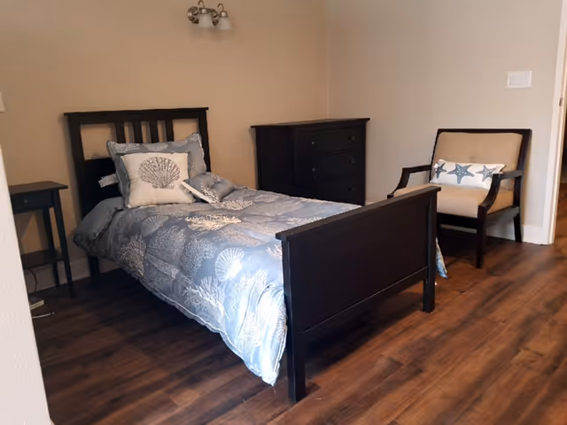 A small bedroom with a single dark wooden bed featuring blue and white seashell-themed bedding and pillows. Next to the bed is a small black nightstand, and against the wall is a dark wooden dresser. A beige armchair with dark wooden arms and legs is positioned near the doorway. The room has wooden flooring and beige walls.