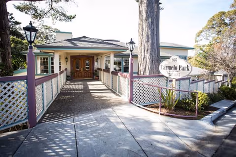 Entrance to Carmelo Park facility with a walkway leading to double wooden doors, flanked by white lattice fencing and two black lamp posts. A large tree stands near the entrance, and a sign reading 'Carmelo Park' is visible on the right side.