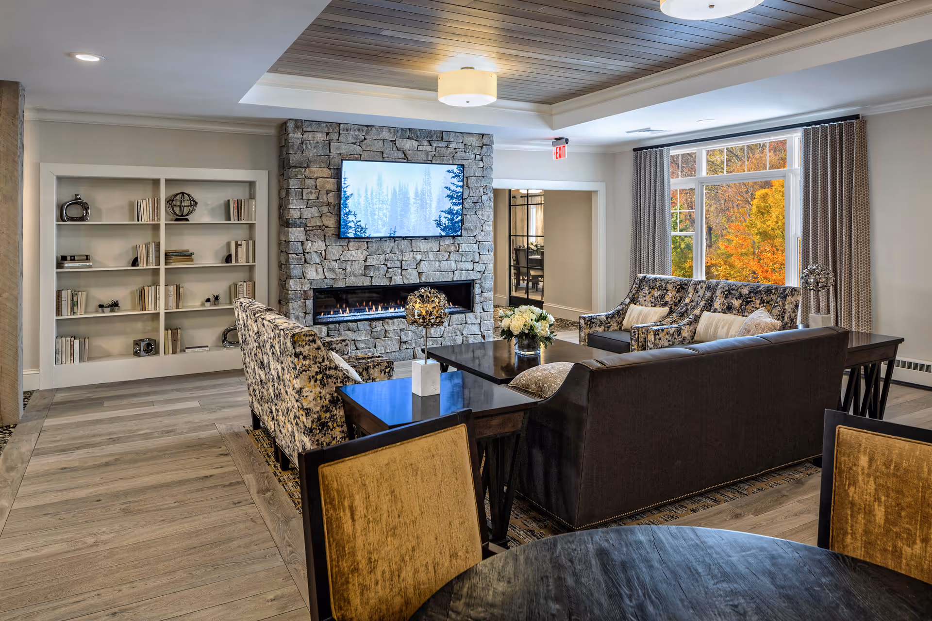 A cozy living room area featuring a stone fireplace with a mounted flat-screen TV above it. The room has a mix of patterned and solid sofas arranged around a dark wooden coffee table with a floral centerpiece. Built-in shelves with decorative items and books are on the left wall. Large windows with curtains reveal colorful autumn trees outside. The ceiling has a recessed wooden panel with a modern light fixture.