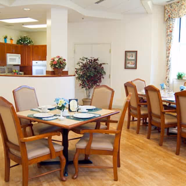A dining area in an assisted living facility with wooden tables and cushioned chairs set with plates, cups, and small flower arrangements. In the background, there is a kitchen area with wooden cabinets, a microwave, and an oven. The room has light-colored walls, a potted plant, framed artwork, and a window with floral curtains.