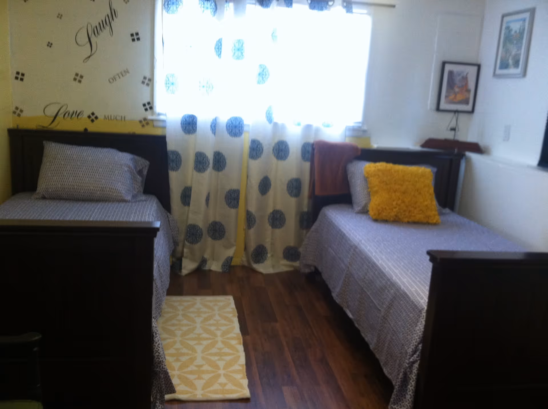 A small bedroom with two single beds placed parallel to each other. Each bed has a patterned bedspread and pillows, one with a yellow decorative pillow. Between the beds is a window with white curtains featuring large blue polka dots. The wall behind one bed has decorative text that reads 'Laugh Often Love Much'. The floor is wooden with a small yellow and white patterned rug between the beds. There are framed pictures on the walls.
