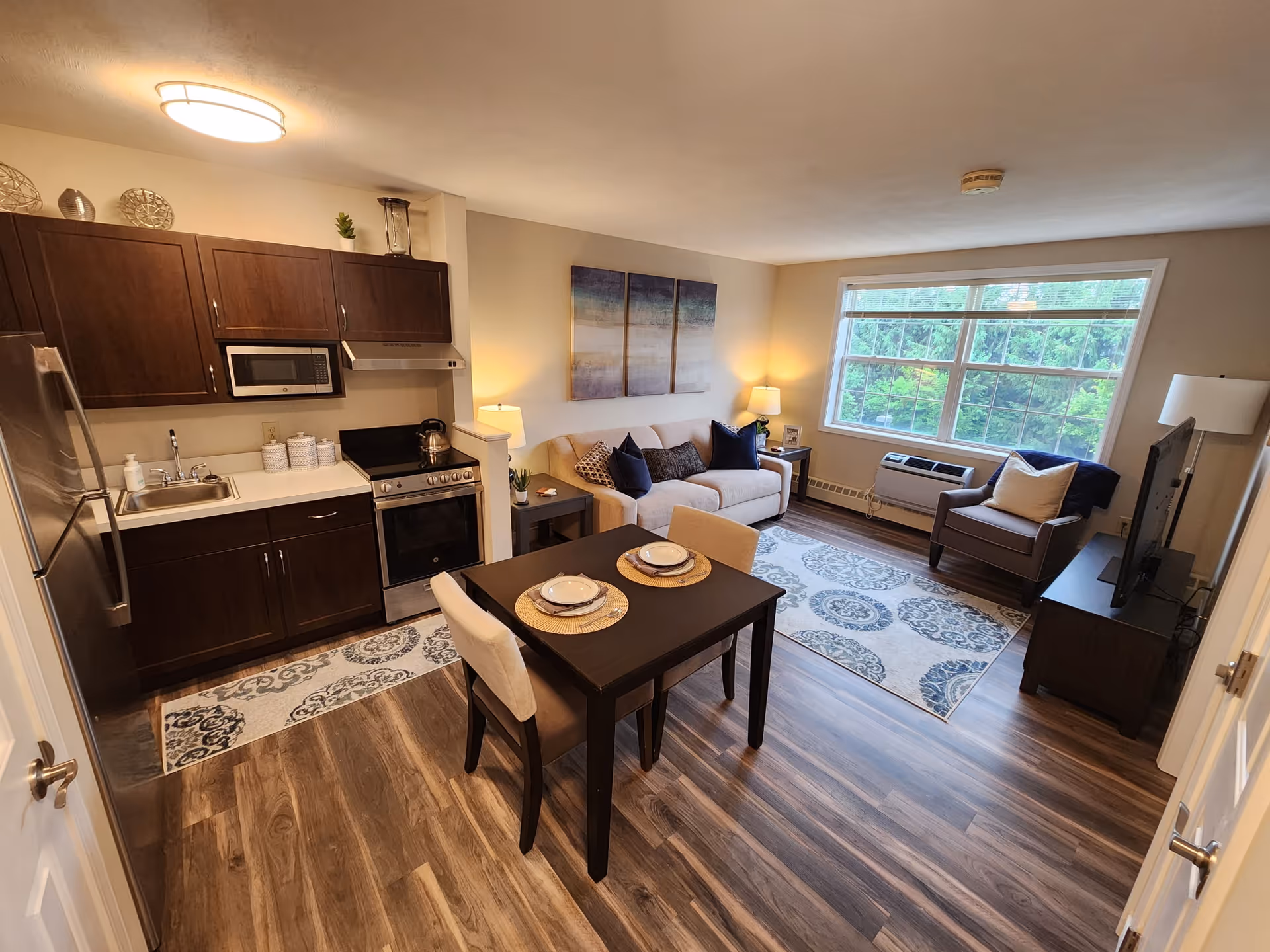 A cozy and well-lit living space featuring a small kitchen area with dark wood cabinets, a stainless steel refrigerator, microwave, stove, and sink. Adjacent to the kitchen is a dining table set for two with plates and placemats. The living area includes a beige sofa with decorative pillows, a side table with a lamp, a patterned area rug, an armchair with a pillow, a TV on a stand, and a large window letting in natural light with a view of greenery outside.