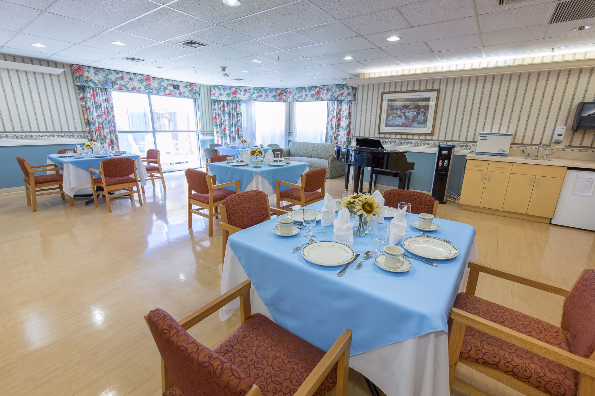 A bright dining room with several tables covered in light blue tablecloths and set with plates, cups, glasses, and napkins. The room has floral curtains, a piano, a couch, and a kitchenette area with cabinets and a sink. The floor is light wood, and the walls have striped wallpaper with a floral border.