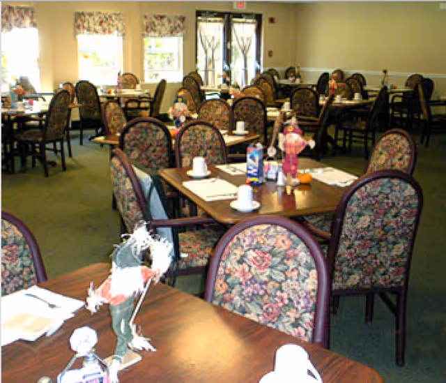 A dining room with multiple tables and chairs featuring floral upholstery. Each table is set with placemats, cups, and decorative figurines. Large windows with floral valances allow natural light into the room.