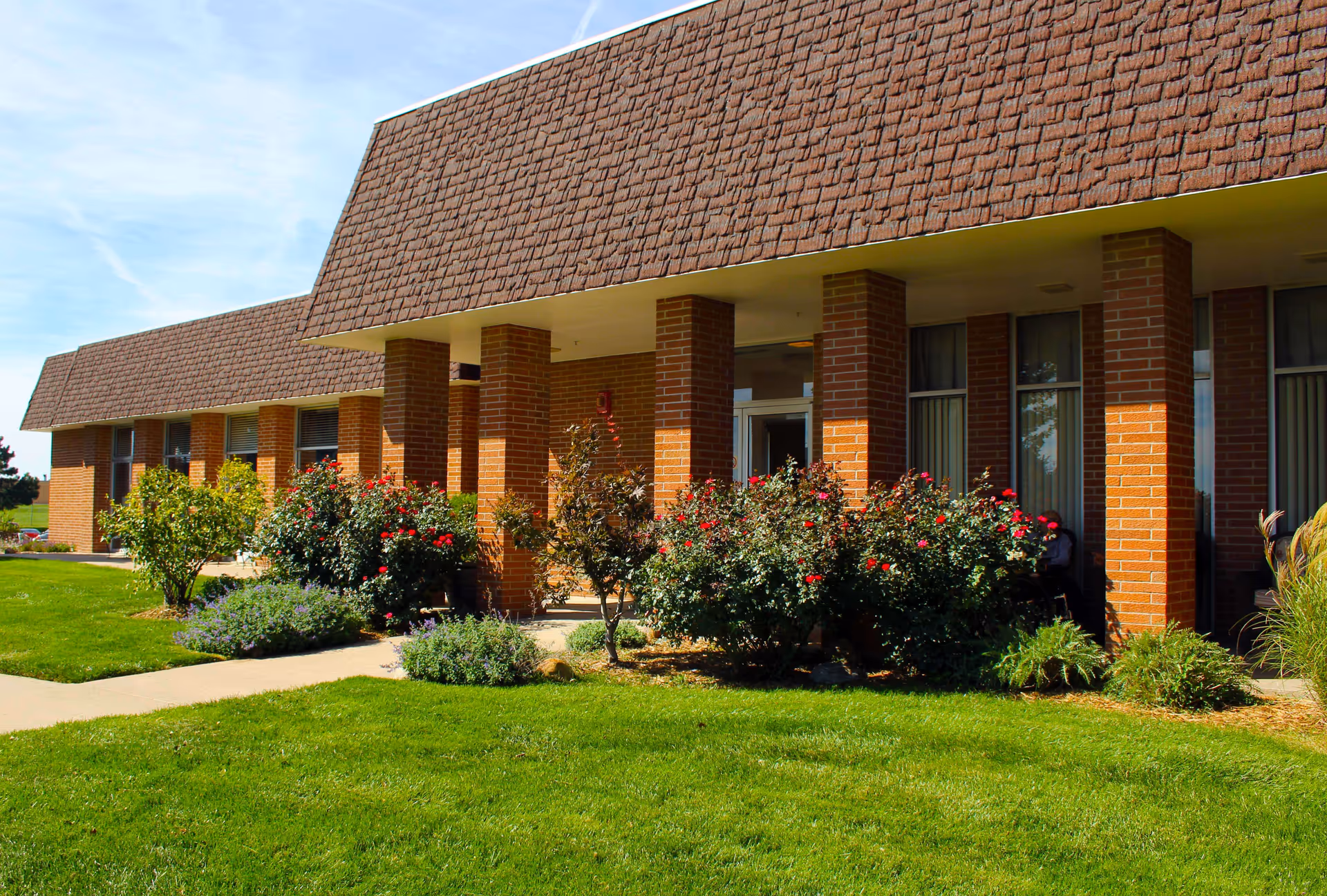 Exterior view of a brick building with a shingled roof, surrounded by well-maintained green grass and flowering bushes under a clear blue sky.