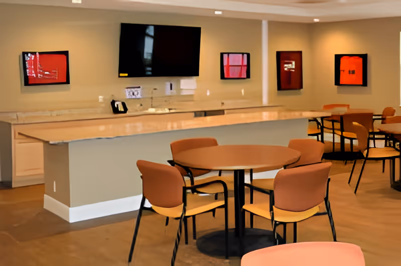 A communal dining area with round tables and chairs, a long counter with a marble countertop, a mounted flat-screen TV on the wall, and four framed red and black artworks hanging on the beige walls.