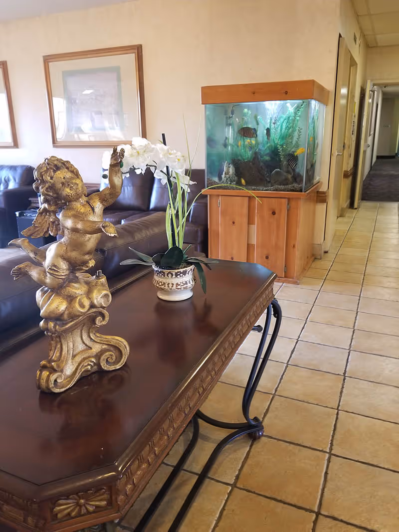 Interior view of a senior living facility lounge area featuring a wooden table with a decorative golden cherub statue and a potted white orchid. In the background, there is a large fish tank with colorful fish on a wooden stand, brown leather sofas, framed artwork on the wall, and a tiled floor leading to a hallway.
