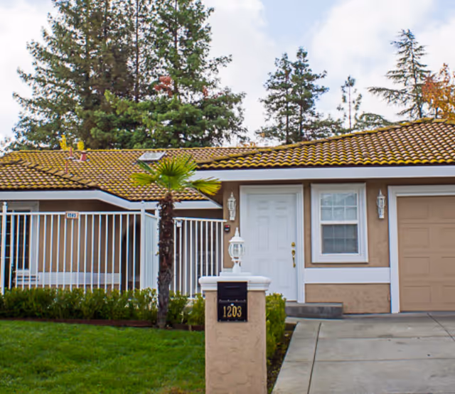 Exterior view of a single-story residential building with a tiled roof, a white front door, a window, a garage door, a small palm tree, and a mailbox with the number 1203. There is a green lawn and some bushes in front of the building, and tall trees in the background under a partly cloudy sky.
