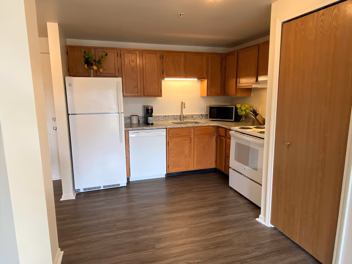 A kitchen area with wooden cabinets, a white refrigerator, dishwasher, stove, microwave, and a sink with a faucet. The floor is dark wood, and there is a small plant with lemons on top of the refrigerator.