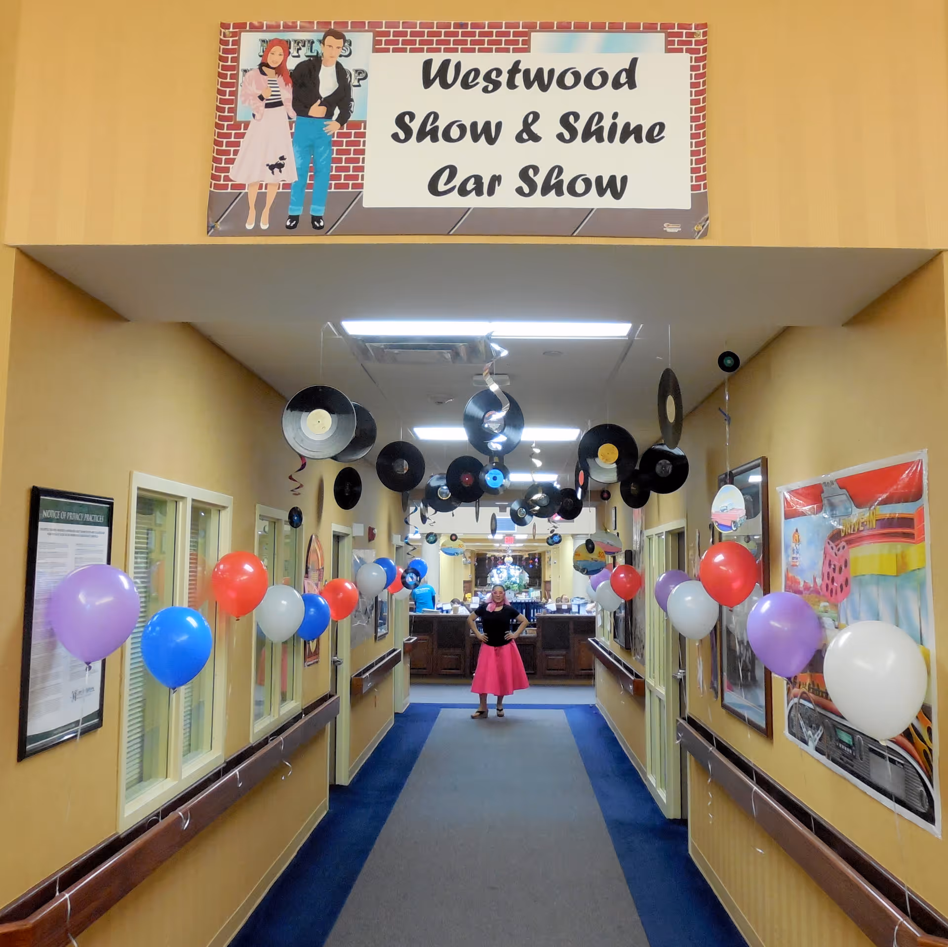 A decorated hallway in Westwood Specialty Care with a banner reading 'Westwood Show & Shine Car Show'. The hallway is adorned with hanging vinyl records and colorful balloons in red, white, blue, and purple. A person dressed in a 1950s style outfit stands in the middle of the hallway with hands on hips. The walls have framed pictures and notices.