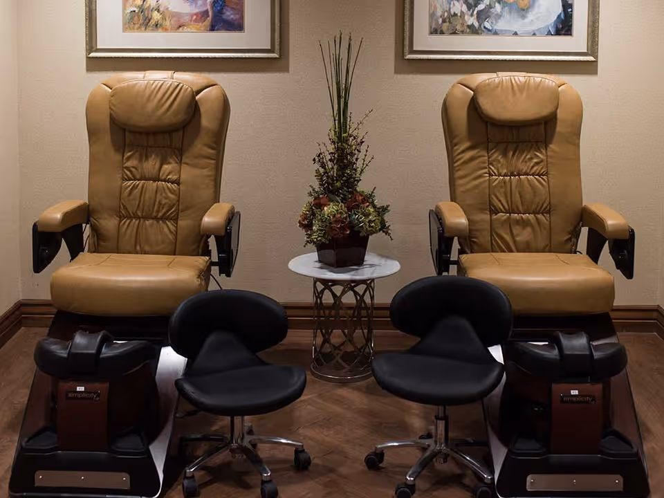 Two tan leather pedicure chairs with black foot baths and black rolling stools in front of them, separated by a small round table with a floral arrangement, set against a beige wall with framed artwork above.