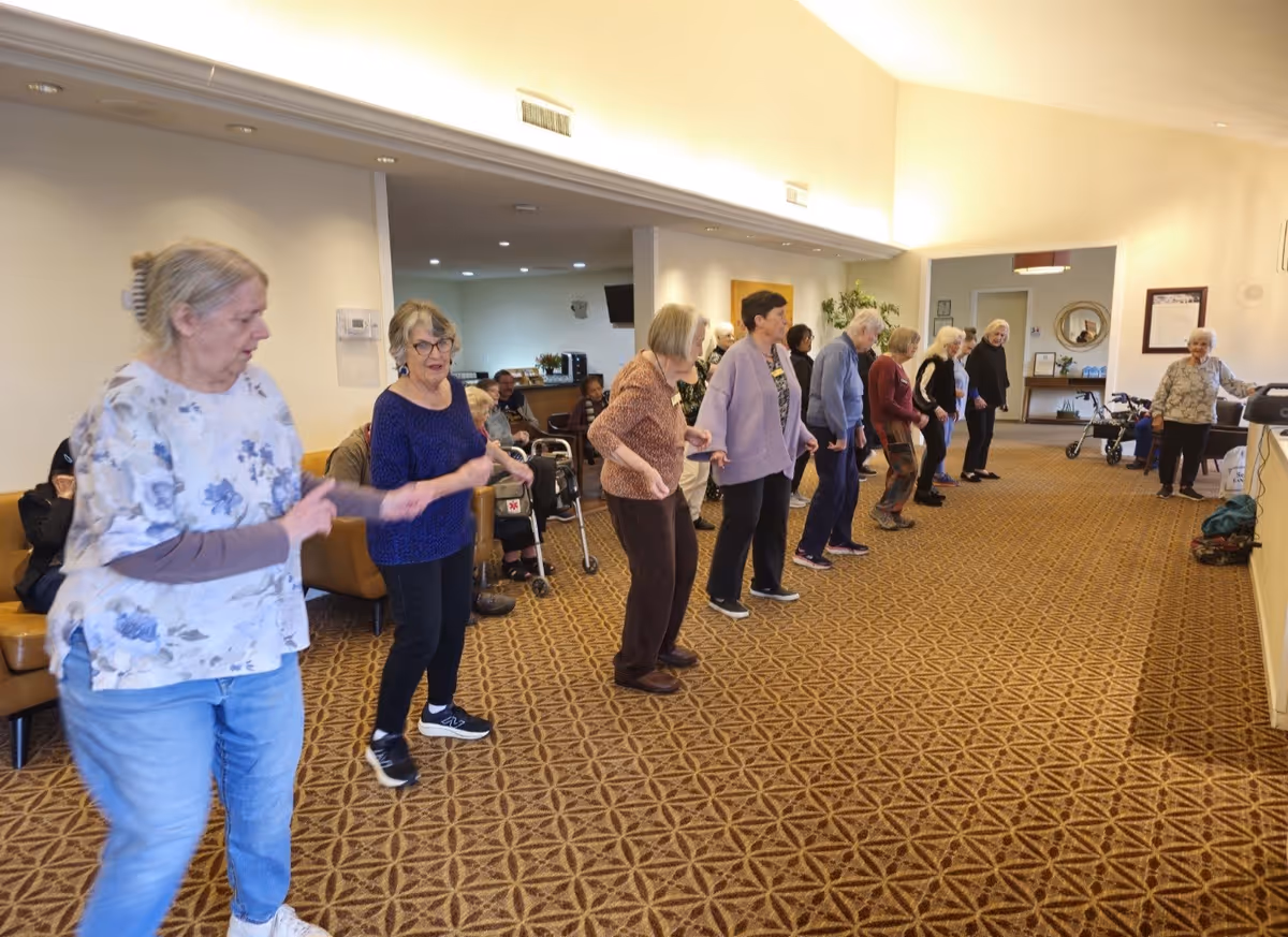 A group of elderly women standing in a line inside a spacious room with patterned carpet, engaging in a group activity or exercise. Some seated individuals and mobility aids are visible in the background, along with neutral-colored walls and ceiling lights.