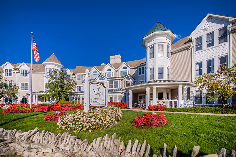 Exterior view of Bickford of Upper Arlington, an assisted living and memory care facility, featuring a large multi-story building with white siding, a turret, and a covered porch. The well-maintained lawn has colorful flower beds, a stone fence in the foreground, and an American flag on a flagpole.