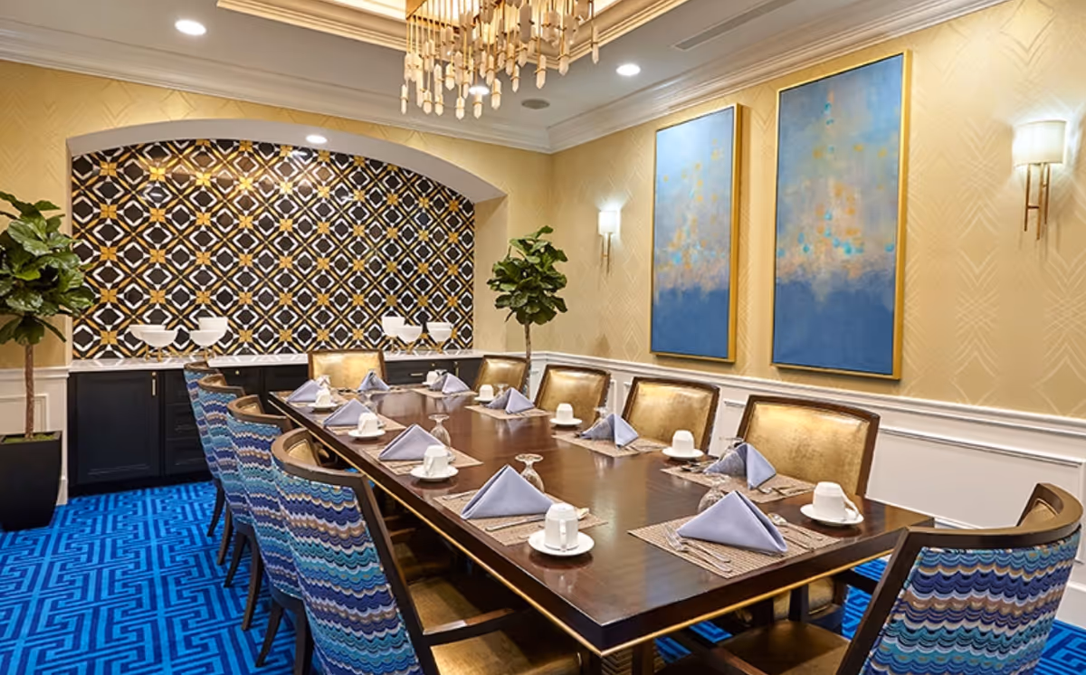 A formal dining room with a long wooden table set for ten people. Each place setting includes a white cup and saucer, a glass turned upside down, and a folded lavender napkin on a woven placemat. The chairs have a blue and purple wave pattern on the backrests and gold upholstery on the seats. The room features a blue patterned carpet, yellow walls with white wainscoting, two large blue and gold abstract paintings, two potted plants, and a decorative chandelier hanging from the ceiling. There is a black and gold geometric patterned alcove with white bowls on a counter at the far end of the room.