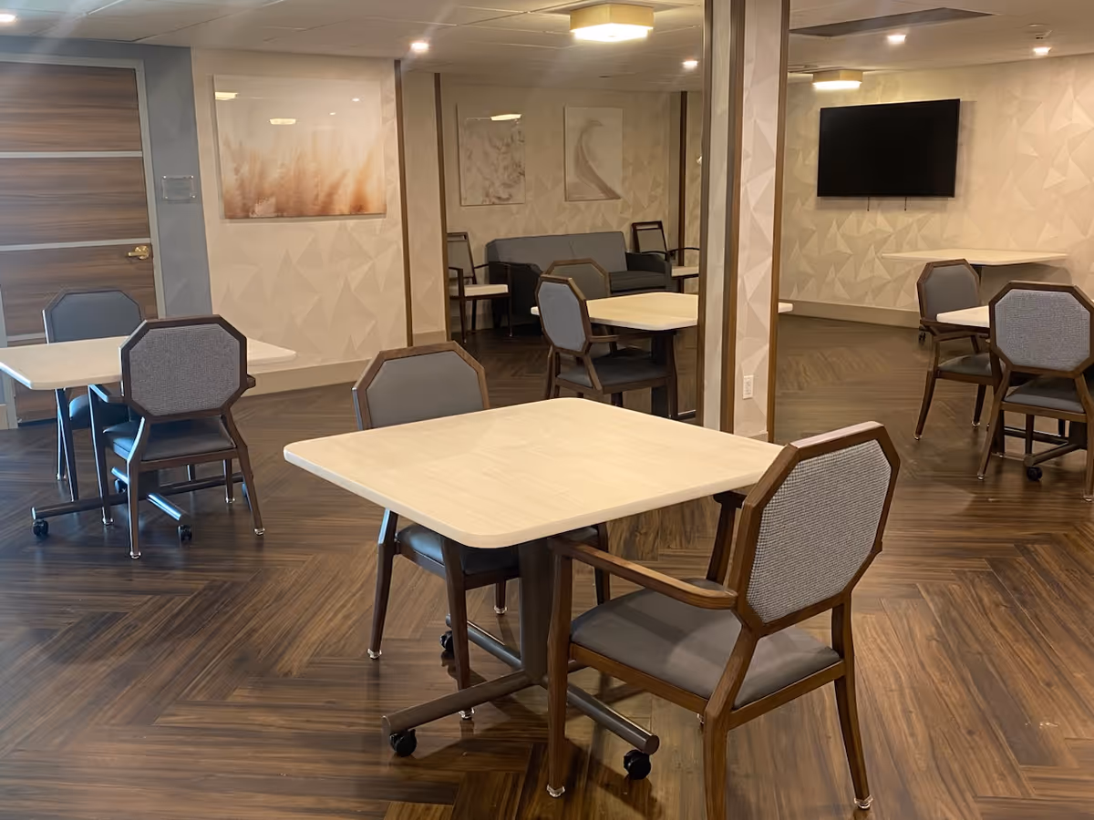 A dining area with several square tables and cushioned chairs on wheels. The room has wood-patterned flooring, light-colored walls with geometric patterns, and framed artwork. A flat-screen TV is mounted on one wall, and there is a seating area with a sofa and chairs in the background.