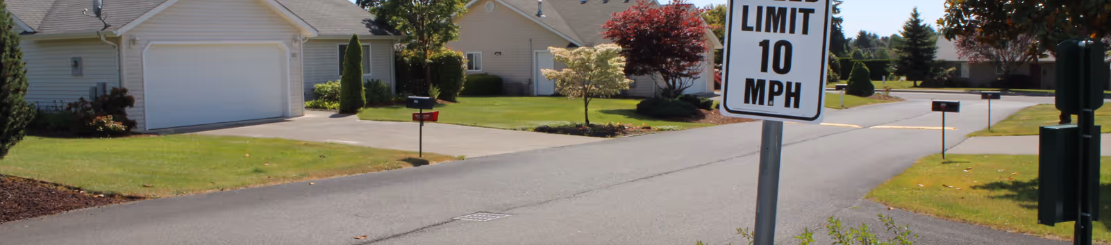 A quiet residential street in a senior living community with single-story houses, well-maintained lawns, small trees, and a speed limit sign indicating 10 MPH.