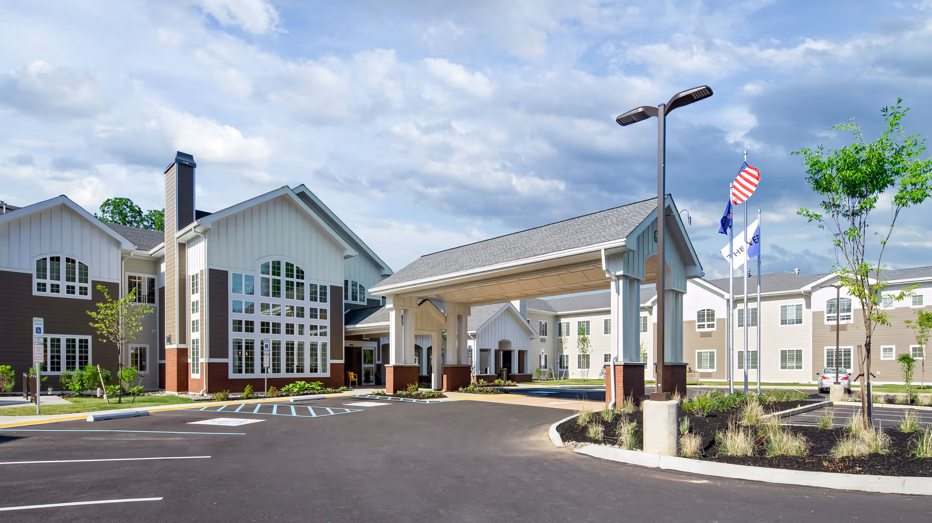 Exterior view of The Vero at Bethlehem senior living facility showing a large covered entrance with brick pillars, multiple windows, a parking lot with handicap spaces, three flagpoles with flags, and landscaped areas with young trees and plants under a partly cloudy sky.