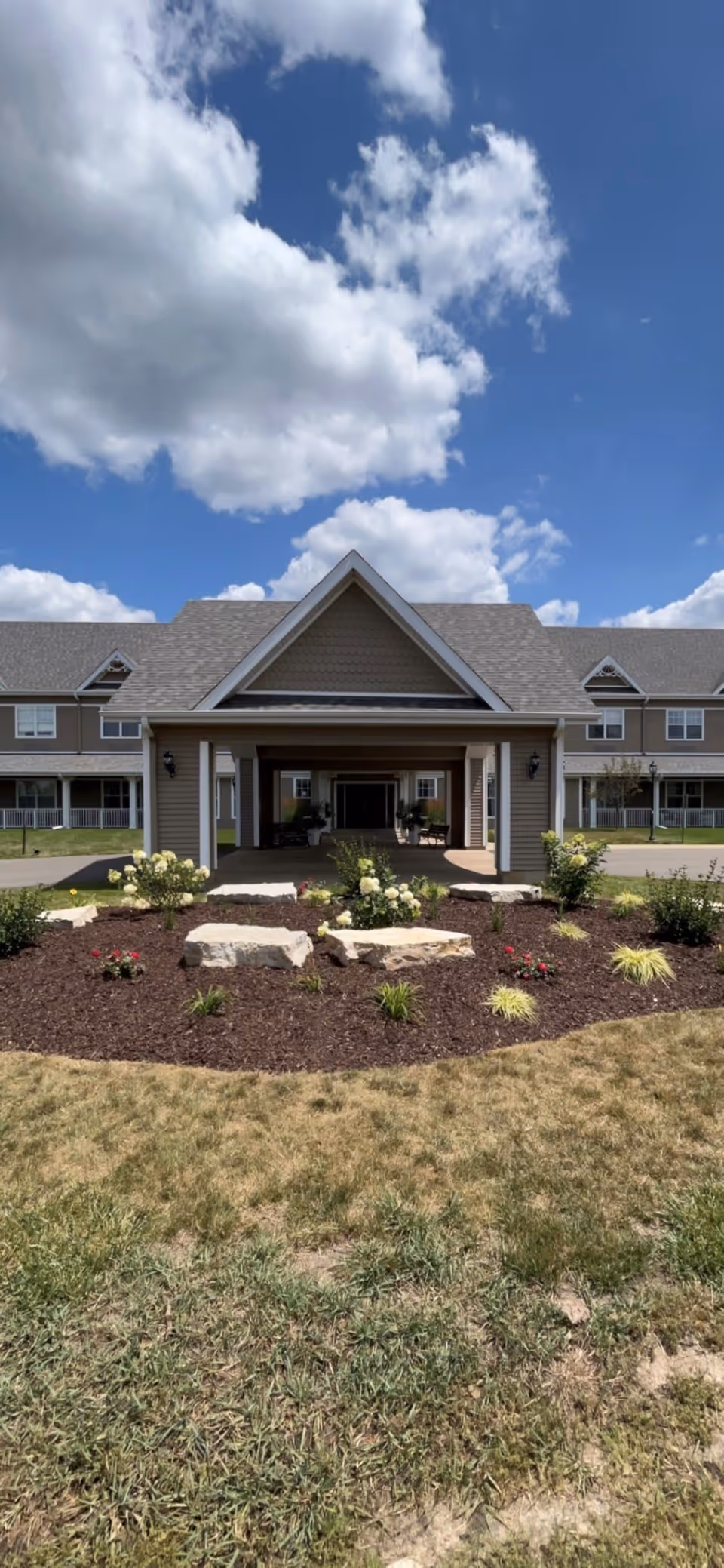 Front exterior view of a senior living facility building with a covered entrance, landscaped garden with flowers and rocks, and a partly cloudy blue sky above.