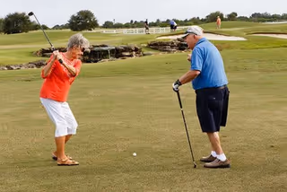 An elderly woman in a red shirt and white pants is preparing to swing a golf club while an elderly man in a blue shirt and black shorts watches on a golf course with green grass and a small waterfall in the background.