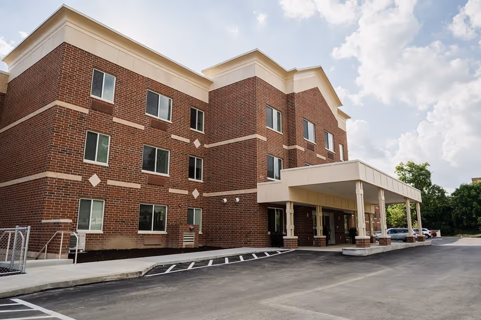 Exterior view of a three-story brick building with multiple windows and a covered entrance area. The building is surrounded by a paved parking lot with marked parking spaces. Trees and a partly cloudy sky are visible in the background.