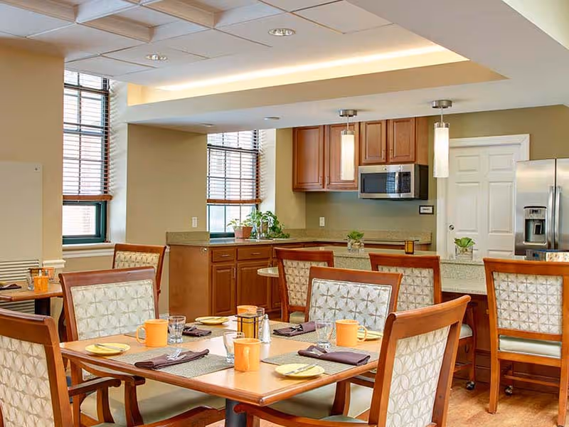 A bright dining area with a wooden table set for four, featuring orange mugs, yellow plates, and brown napkins. The room has wooden chairs with patterned cushions, large windows with blinds, and a kitchen area in the background with wooden cabinets, a microwave, and a refrigerator. There are also some small plants on the counter.