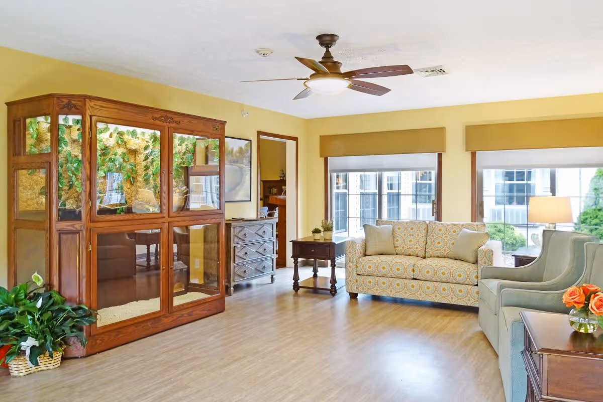 A bright living room with light yellow walls and wood flooring. The room features a patterned sofa, a light blue armchair, a wooden side table with a vase of orange flowers, and a ceiling fan with a light. There is a large wooden cabinet with glass panels and green plants inside, and large windows with beige valances letting in natural light.