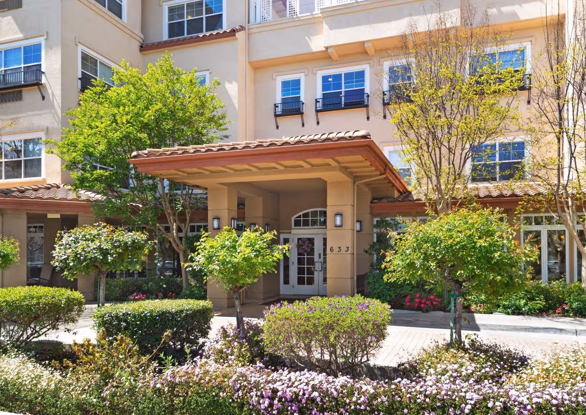 Exterior view of a senior living facility entrance with a covered porch, surrounded by well-maintained bushes, flowering plants, and trees. The building has multiple windows with black railings and beige walls with a tiled roof.