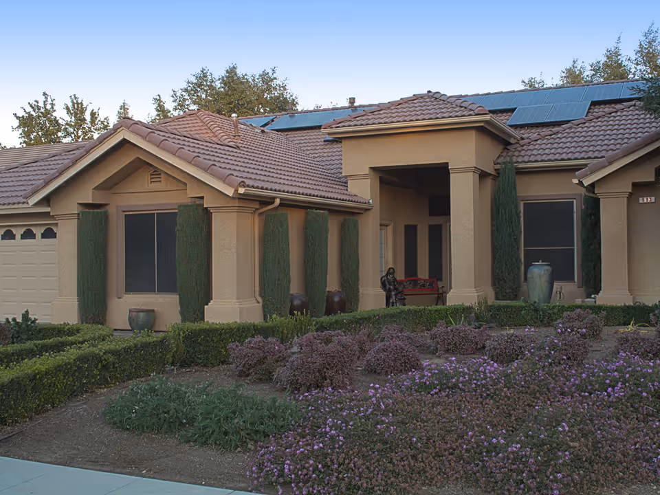 Exterior view of a single-story residential building with a tiled roof and solar panels. The entrance features a covered porch with columns, a red bench, and a statue. The front yard is landscaped with trimmed bushes, tall narrow trees, and flowering plants.