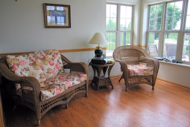 A cozy sitting area with wicker furniture including a floral cushioned loveseat and armchair, a small wicker side table with a lamp, and large windows letting in natural light overlooking a green outdoor space.
