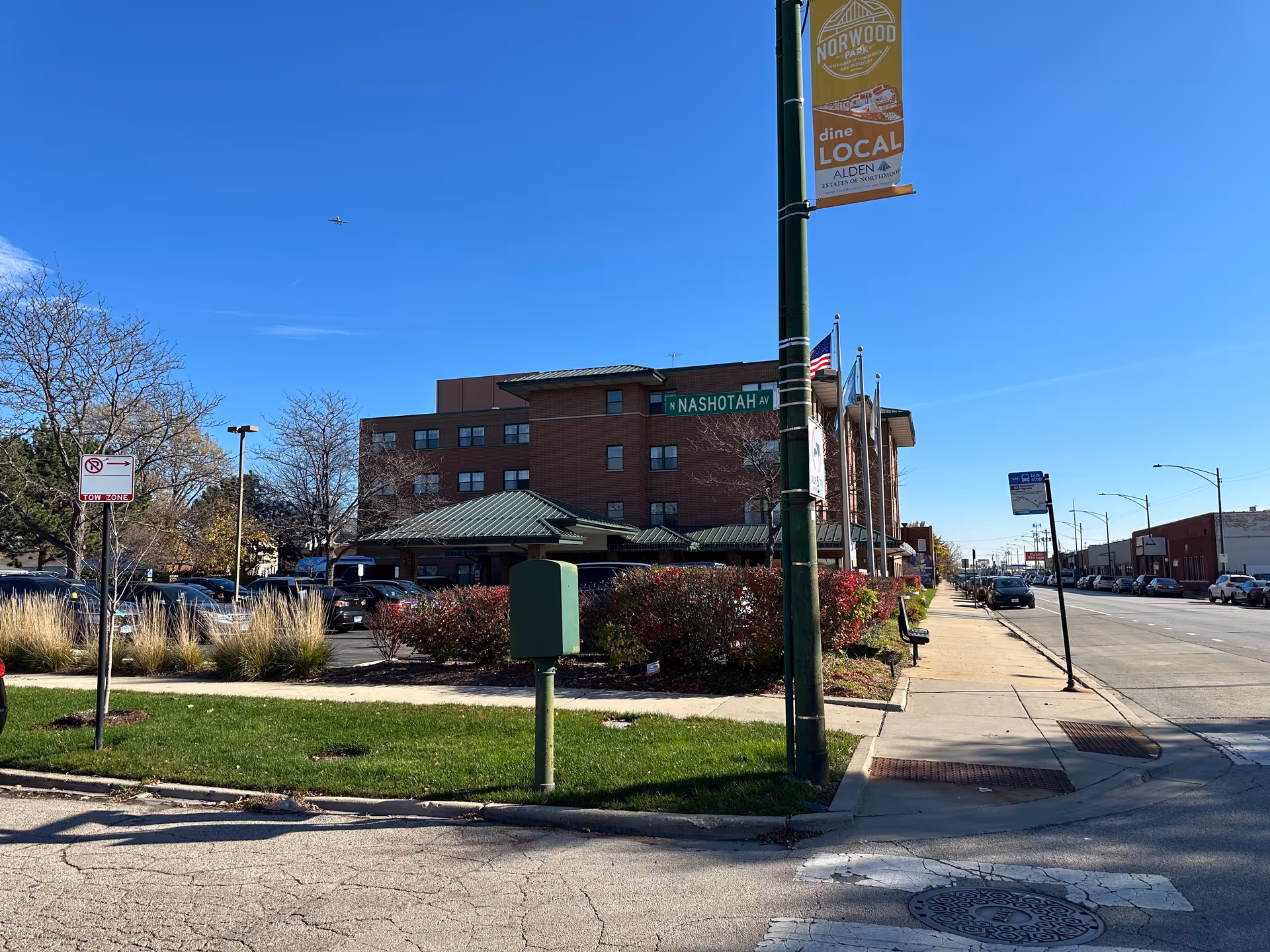 Exterior view of Alden Estates Of Northmoor, a multi-story brick building with a green roof, located at the corner of N Nashotah Avenue. The scene includes a street with parked cars, a bus stop sign, a green mailbox, and a street lamp with a banner promoting Norwood Park and Alden Estates Of Northmoor. The sky is clear and blue.