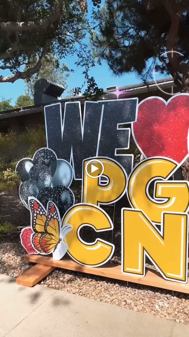 Outdoor decorative sign with large letters spelling out 'WE ❤️ PGCN' with a butterfly and balloons, set against a backdrop of trees and a building under a clear blue sky.