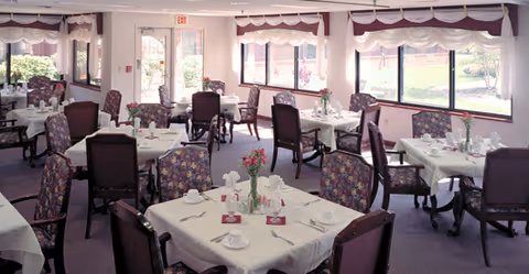 A bright dining room with multiple tables covered in white tablecloths, each set with cups, saucers, napkins, and small flower arrangements. The room has large windows letting in natural light and floral-patterned chairs around the tables.