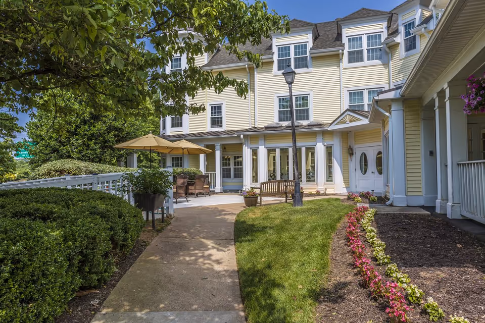 Outdoor patio area of a senior living facility with a paved walkway, green bushes, a flower bed with pink and white flowers, a wooden bench, patio tables with umbrellas, and a yellow multi-story building in the background under a clear blue sky.