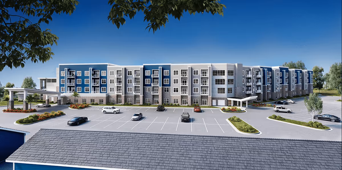 Exterior view of a modern multi-story senior living facility building with blue and gray accents, multiple windows, balconies, and a large parking lot in front. The sky is clear and blue, and there are trees and landscaped areas around the building.